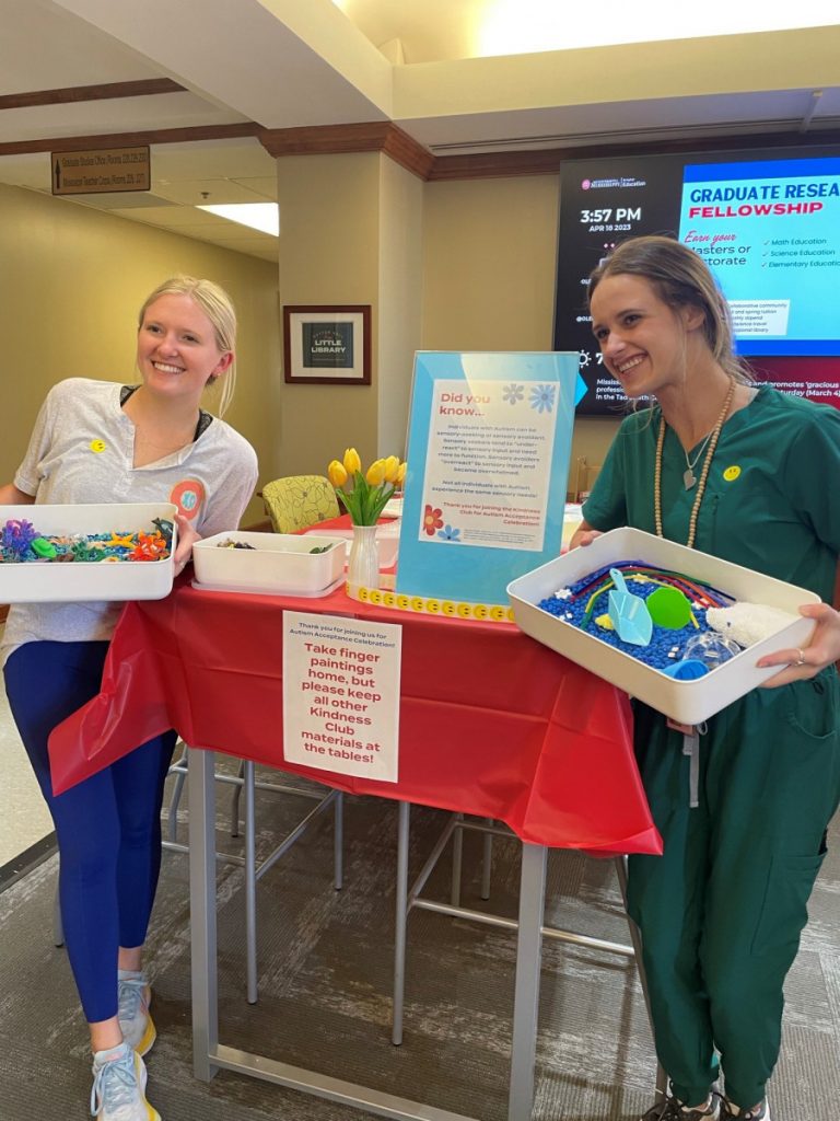 Kindness Club President Abby Newton (left) and Secretary Kennedy Robinson set up for the Autism Acceptance Celebration. The two are holding up sensory kits to show the colorful and textured contents.
