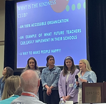 The Kindness Club presents at the Equity in Education Summit, inviting teachers to participate in National Bus Driver Appreciation Day. Four group members stand behind a podium.
