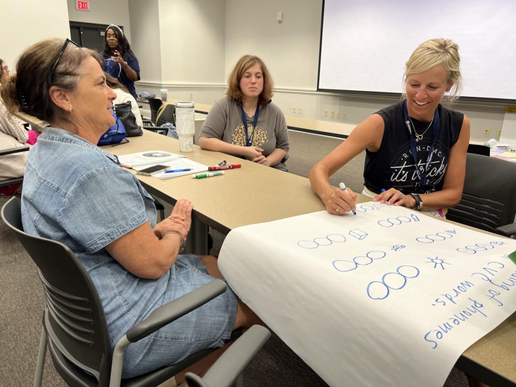 Three smiling  teachers on either side of a table map ideas with a blue marker on a large roll of paper.